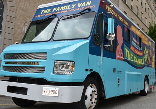 A brightly painted blue mobile clinic van parked on a city street. The side reads “The Family Van” and advertises free health screenings, with the website and phone number displayed above the windshield.