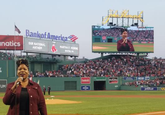 Martha Vedrine singing on the field at Fenway Park, with her image displayed on the jumbotron in the background