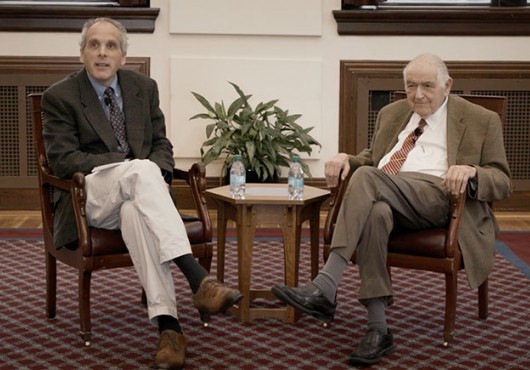 Two men in sports coats and ties sit in armchairs in front of a small table.