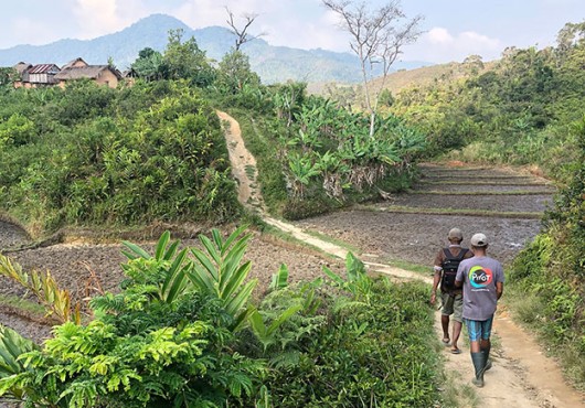 Two people walk along a dirt path bordered by green vegetation and rice paddies in a hilly rural area.