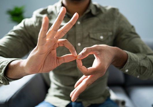 Close-up of two hands belonging to a person of color as they form the ASL sign for “interpreter.” The person’s head is unseen as they sit in a chair in a button-down shirt.