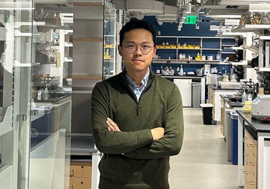 A man stands in a chemistry lab, with machinery on lab benches and reflections on glass partitions. 