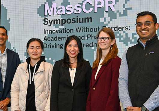 Five people in business clothes pose for a photo in front of a screen with the MassCPR symposium name