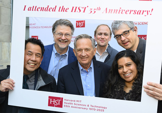 Six people pose, smiling, inside a large portable photo frame that says “Harvard-MIT Health Sciences & Technology 55th Anniversary 1970-2025”