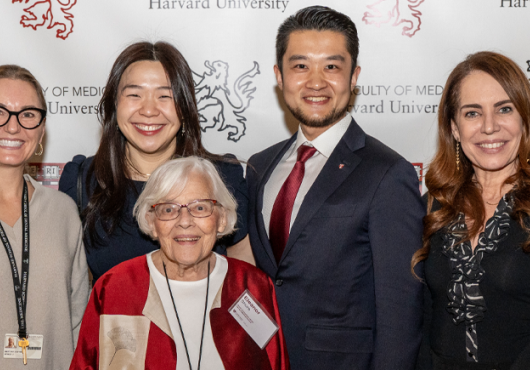 A group of adults stands in front of a backdrop displaying the Harvard University Faculty of Medicine logo. They are smiling and posing for a group photo. The group includes an older woman in a red and beige jacket standing in front. Everyone is dressed in professional or semiformal attire.