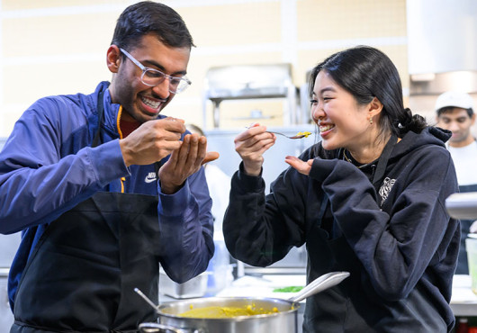 Two young adults wearing aprons taste a spoonful of food from a large pot.