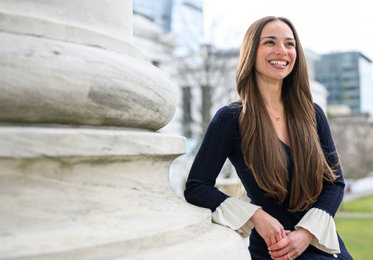 Nikki Pérez smiling, leaning against the base of a column in front of Gordon Hall with the HMS Quad in the background