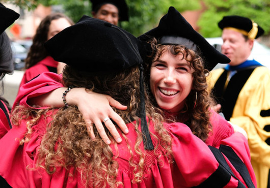 Two women hug and smile in center of photo with other grads and faculty gathered in background.