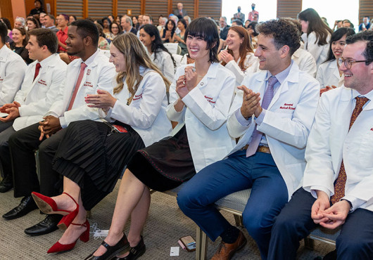 Dozens of students and others sit in rows in a conference room, wearing white coats embroidered with “Harvard Medical School,” smiling and applauding
