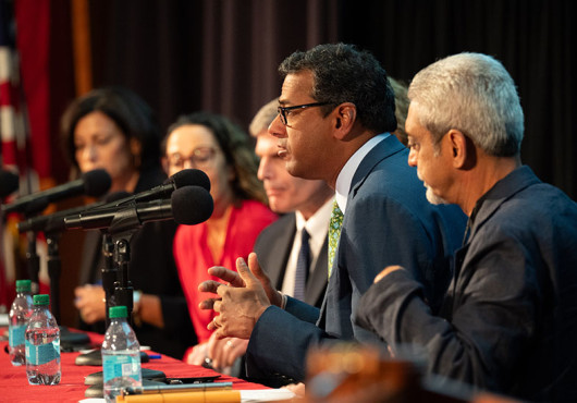 Atul Gawande leans toward a microphone at a long table with four other panelists visible.