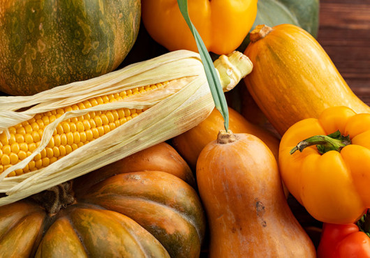 A variety of edible squashes on a wooden table along with bell peppers and an open ear of corn.