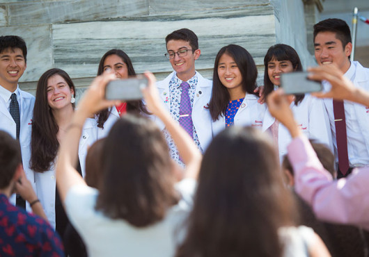 Seven young adults in white coats face the camera smiling, while four adults with their backs to the camera take pictures with their phones.