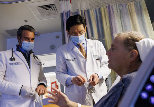 A patient in a hospital bed talks to two surgeons in doctors’ coats and masks.