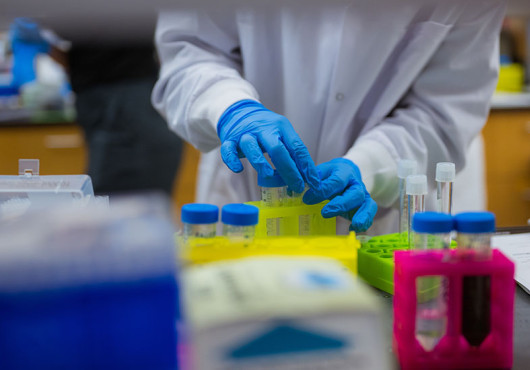 : Scientist wearing a white coat and blue gloves working in a lab setting.