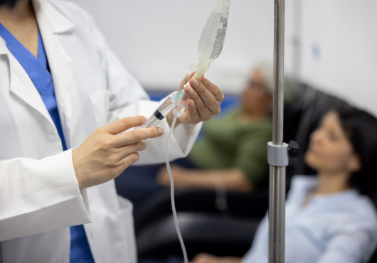 A health care professional administers a medication in a syringe connected to a hanging IV bag beside a seated patient.