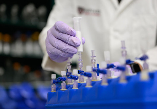A researcher wearing purple gloves handles a syringe connected to laboratory equipment with multiple valves and tubes arranged in a blue rack