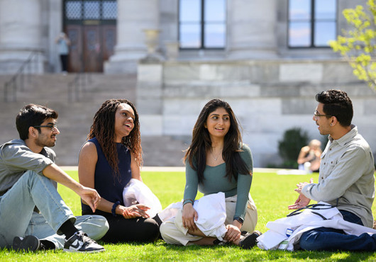 Four young people sit on the grass talking and holding white coats in front of a marble building.