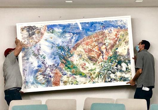 Two men hold a large framed piece of art at an angle while trying to attach it to four hooks on a plain wall