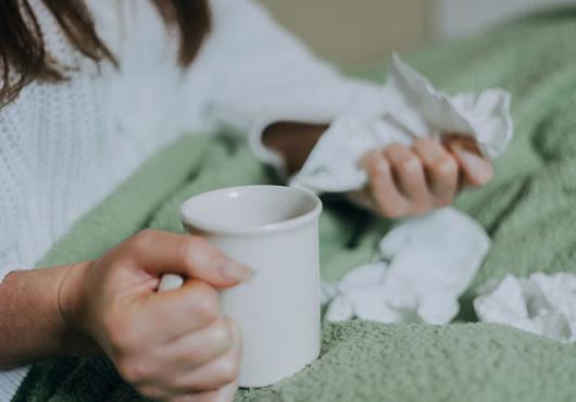 a person sitting in bed under a blanket, holding tea and surrounded by crumpled facial tissues