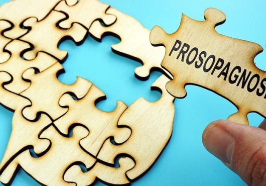 A hand holds a puzzle piece that reads "prosopagnosia" above a puzzle in the shape of a human brain