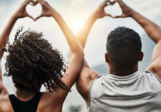 Photo of a Black young man and young woman making heart signs with their hands, backs facing the camera