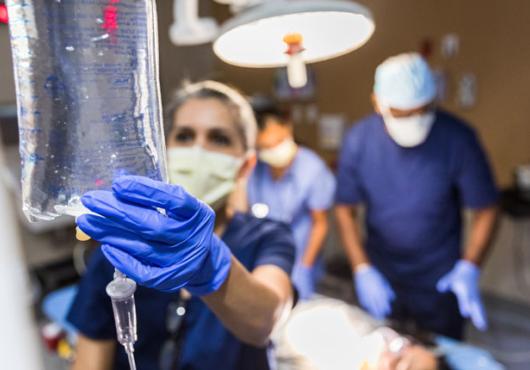 Out-of-focus photo of ER nurse checking an IV bag with a surgeon looking at a patient on a gurney behind her