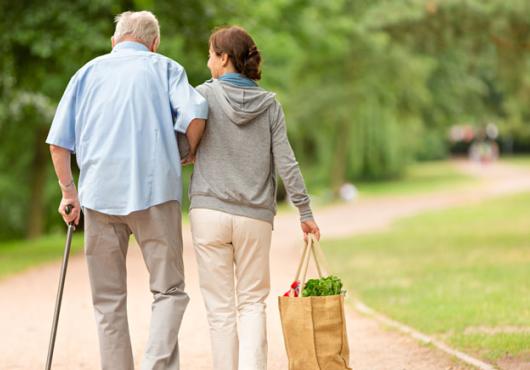 Back view of a woman walking with an older man supported by a cane along a dirt path with trees and grass surrounding