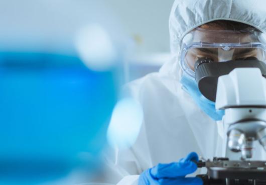 Researcher in white gown and goggles looking in to a microscope