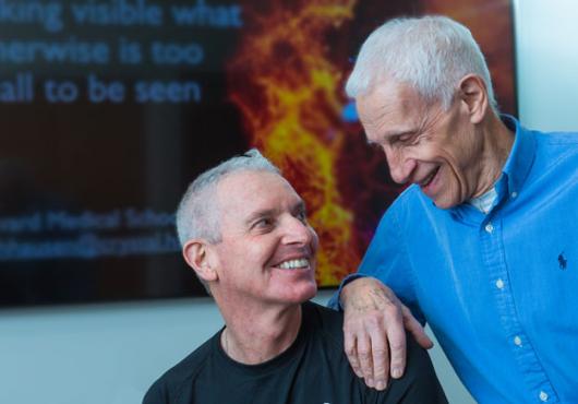 Harrison and Kirchhausen in front of a digital screen with molecular imagery looking into each other's eyes