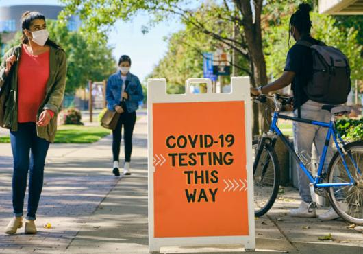 An orange sign that says "COVID-19 testing this way" on a college campus with students walking nearby