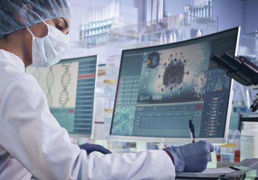A scientist sits writing notes in front of a computer monitor showing a COVID-19 virus