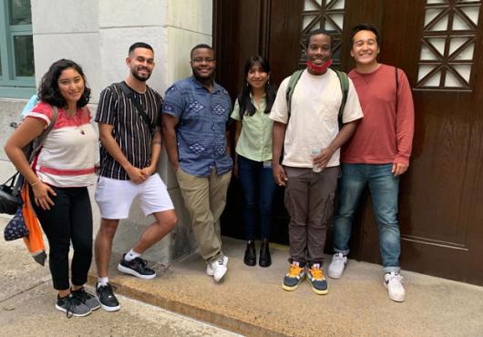 Six students pose for a photo against a stone building and a wooden door