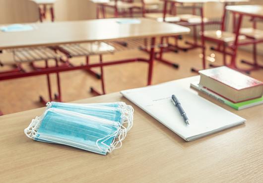 A stack of surgical masks, a pad of paper and pen, and a pile of books sit on a table in an empty classroom