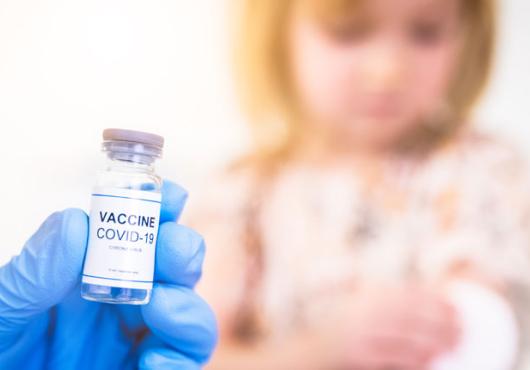 Blue-gloved hand holding COVID vaccine vial and blurry child in background	 