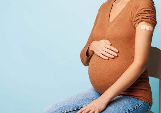 Cropped stock image of a woman with light brown skin sitting in a hair with a hand on her pregnant belly. A bandage on her arm indicates a recent COVID vaccination.