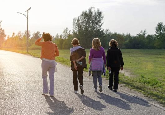 Four adult women walking side-by-side on a path surrounded by green grass