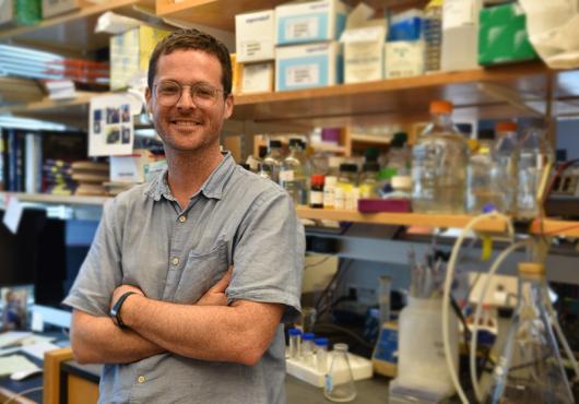 Young white man stands smiling with arms crossed in front of lab bench full of bottles