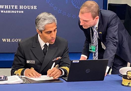 Murthy in uniform seated at table looking at laptop with Beckman to right leaning in; a backdrop behind them reads The White House