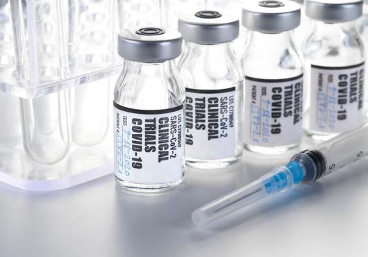 vials labeled "clinical trials COVID 19" in front of test tube rack, with syringe in foreground