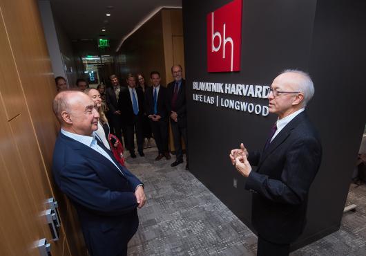 Two men in suits stand in front of a sign with a red square logo with letter B and H above white type on black background that reads Blavatnik Harvard Life Lab Longwood