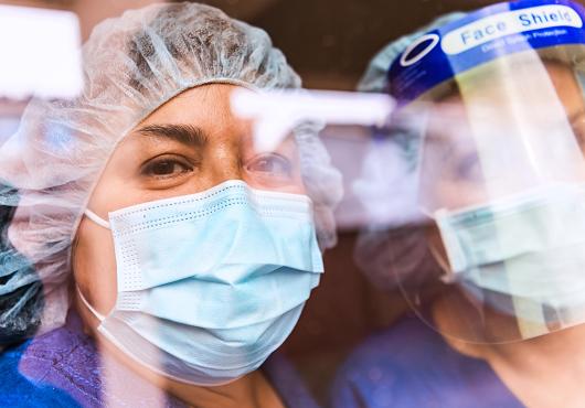 The faces of two people in medical scrubs, protective masks, and head gear seen through a reflection-covered window.