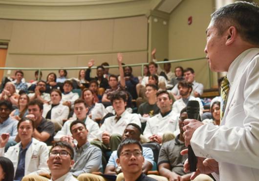 Sideview of Chang in white coat holding mic while speaking to audience of students, many in white coats