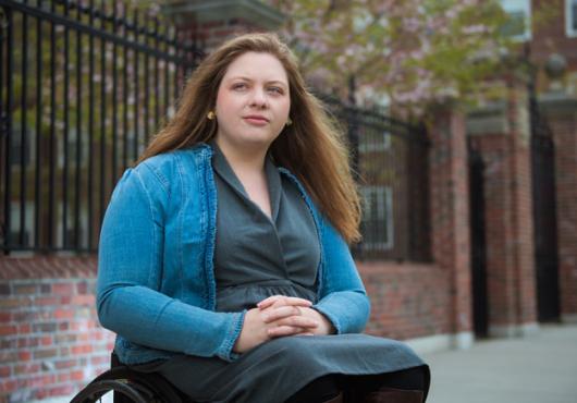 Cloutier sitting in wheel chair with arms folded in lap wearing a green button down and aqua sweater
