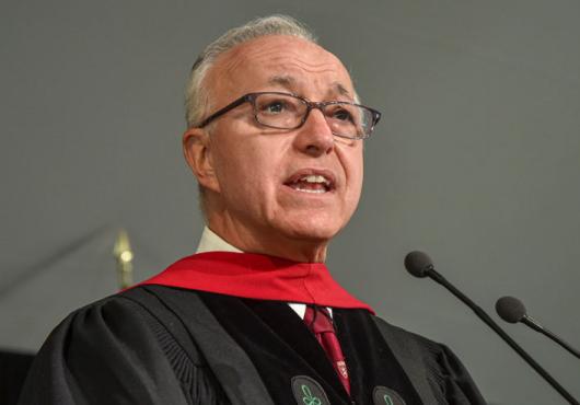 A gray-haired man with glasses and doctoral robes speaks into a microphone