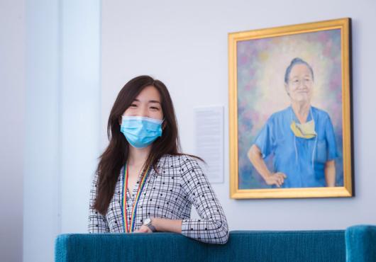 Photo of Pamela Chen, a young Asian woman wearing a mask and patterned shirt standing proudly in front of the portrait she painted of Yeu-Tsu Margaret Lee, who is an older Asian woman in the portrait looking determined while wearing her hair in a bun, scrubs, and a medical mask around her neck
