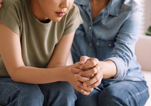 An older person holds a distressed younger person’s hands and shoulder, offering comfort.