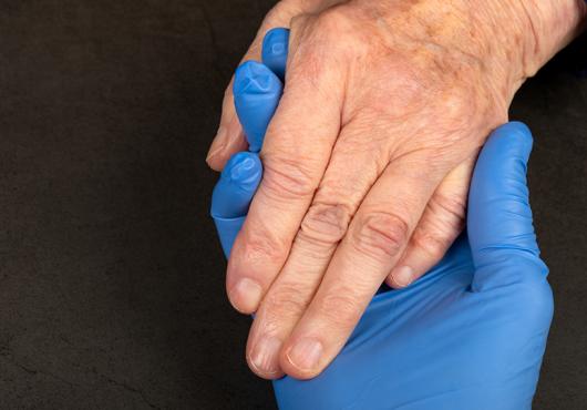 A hand in a blue latex glove holds the ungloved hand of an older white person