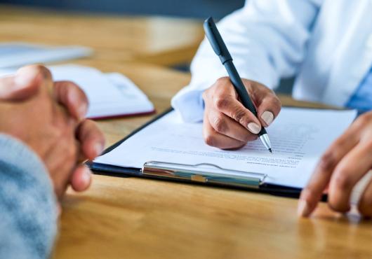 Photograph of doctor's hand writing on a form across from patient with hands folded