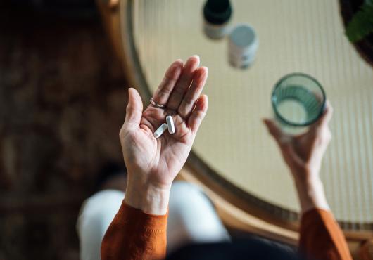 Photo looking down to a woman's hands, one holding a glass of water, in the other hand, some pills