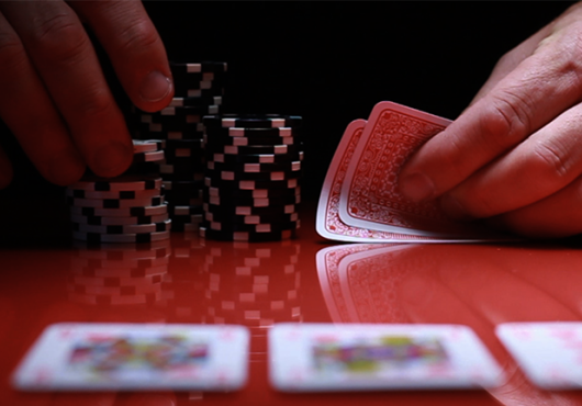 an unseen person at a shiny gambling table holds unknown cards while picking up a few poker chips from a stack. on the table lie three blurry cards face-up.